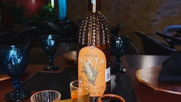 Uma mesa de madeira com uma garrafa de Nîxta, um copo com líquido, um copo shot, paus de canela e uma pequena colher de madeira.