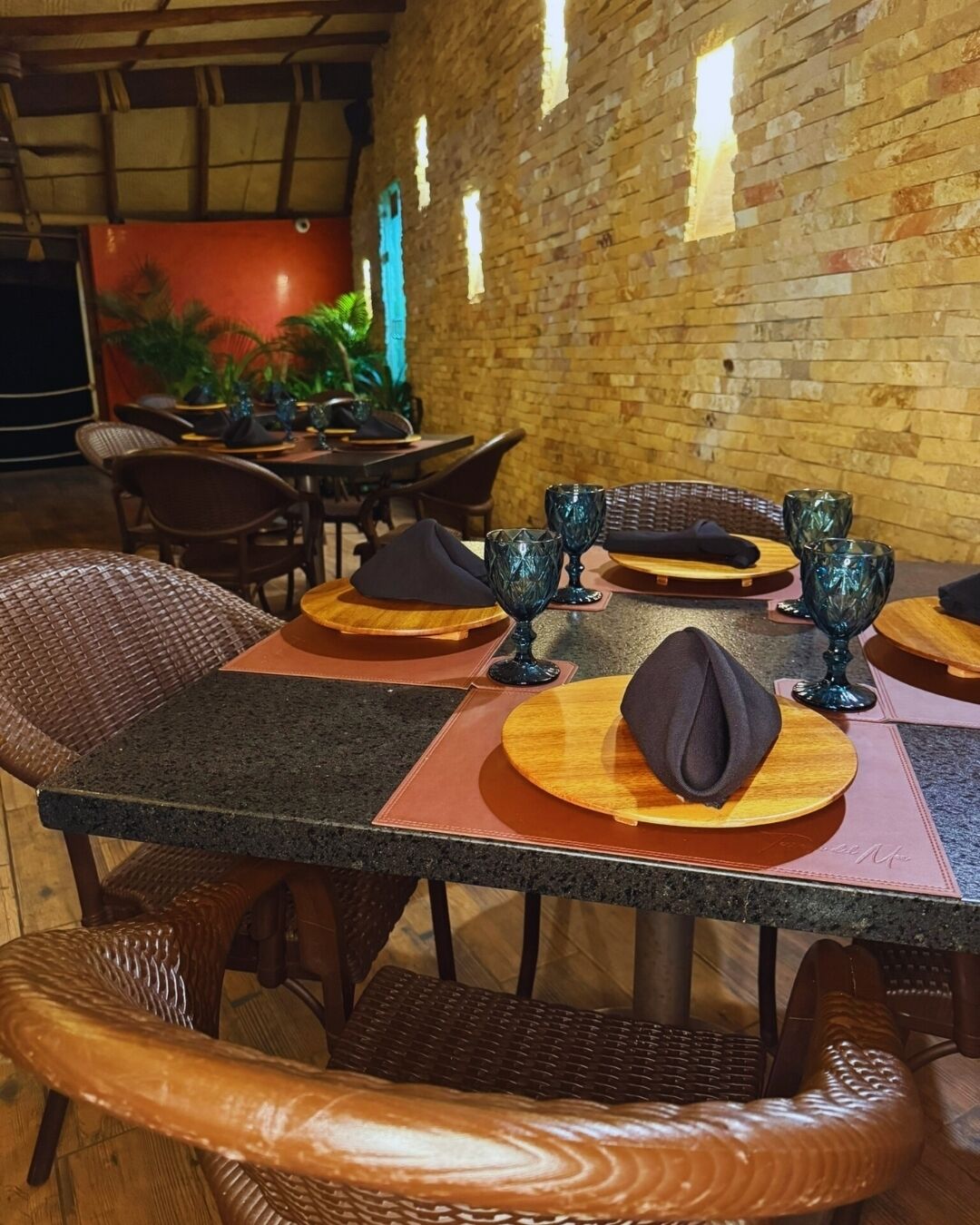 Un espace repas avec un mur en briques, des tables en bois, des chaises en osier et de la verrerie bleue.