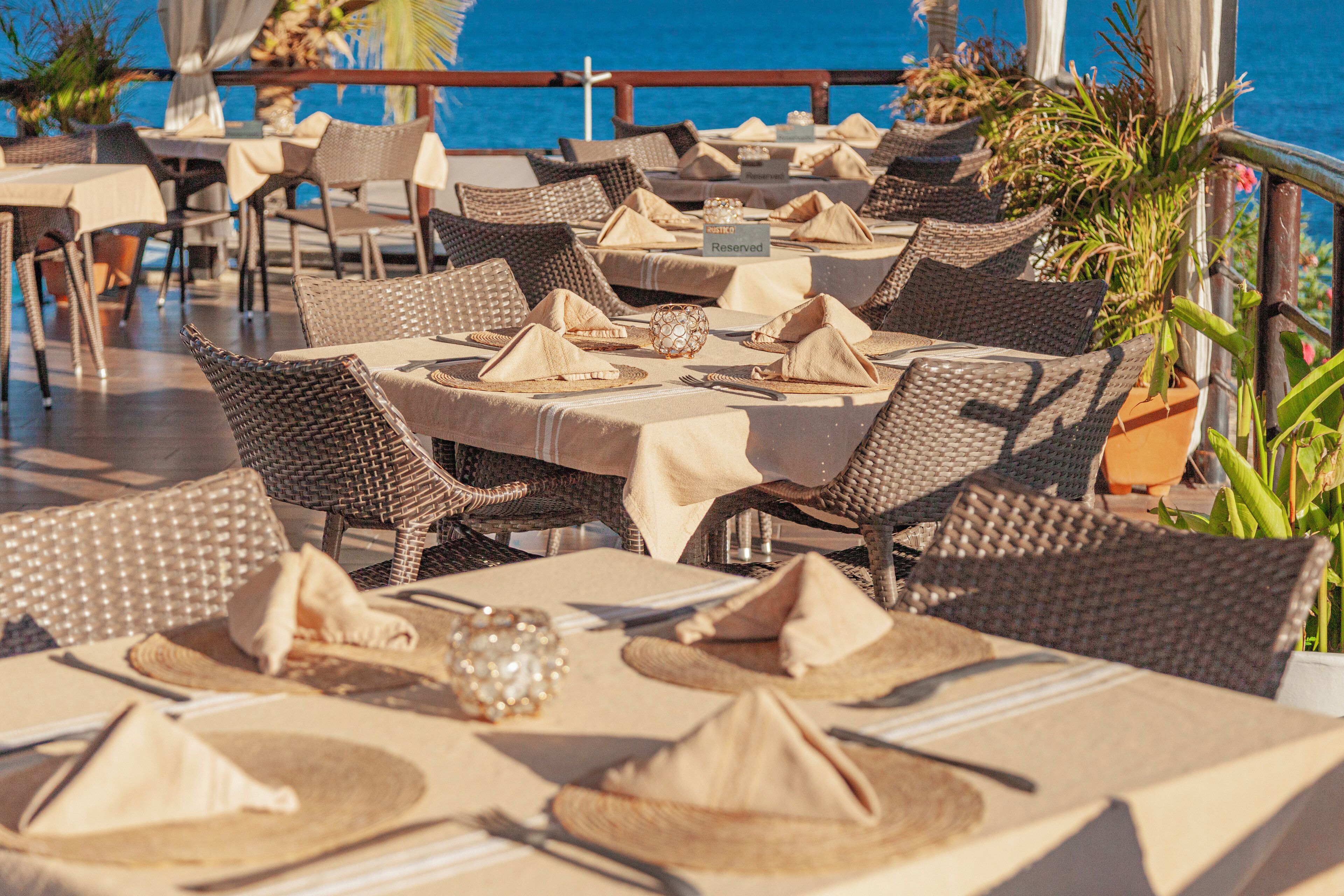 A restaurant with tables set for dining, wicker chairs, and a view of the sea.