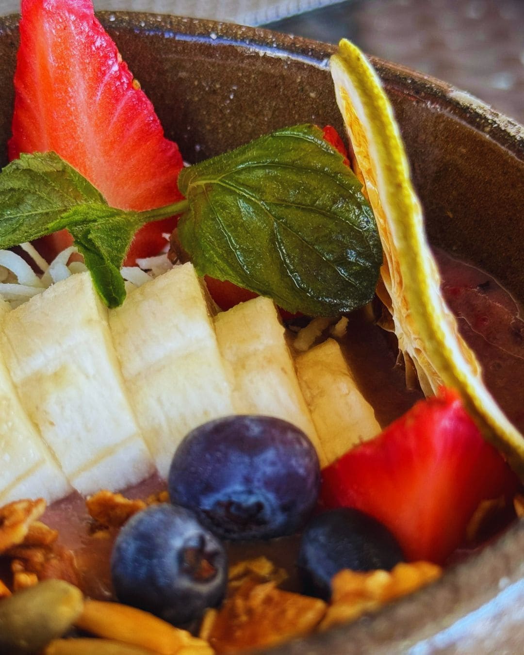 A bowl containing sliced bananas, strawberries, blueberries, and a basil leaf.