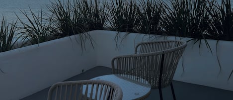 A terrace with two wicker chairs and a table, overlooking a body of water and distant hills.