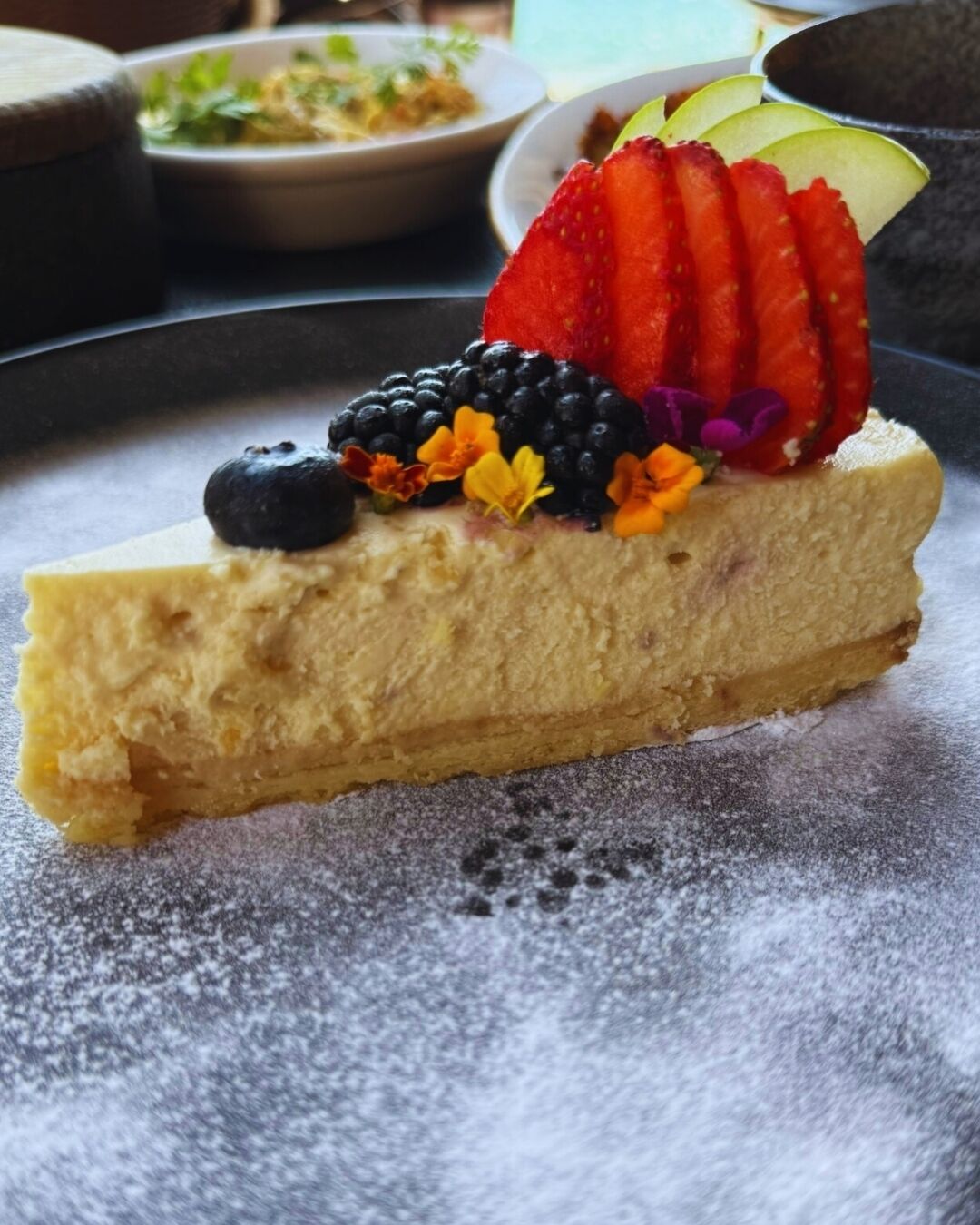 A slice of cheesecake topped with strawberries, blueberries, blackberries, and edible flowers, dusted with powdered sugar.