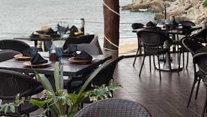 An outdoor dining area with tables and chairs, a thatched roof, and a view of the sea.