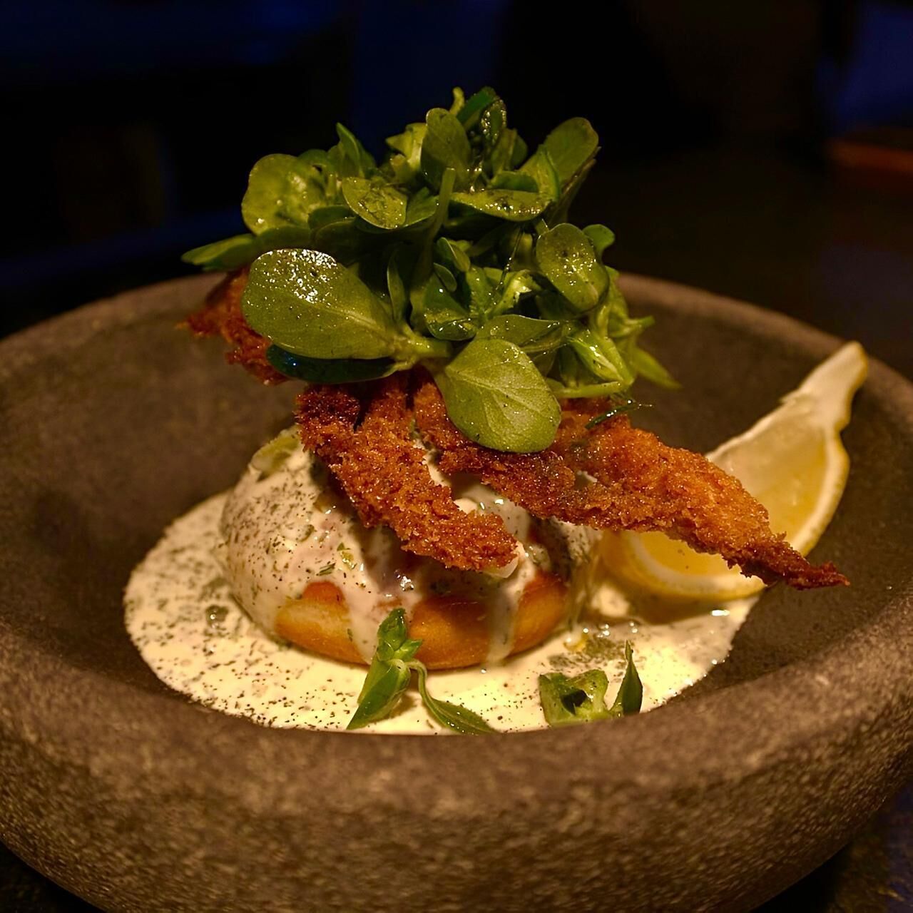 A dish with crispy fried elements, greens, and a lemon wedge on a stone plate.
