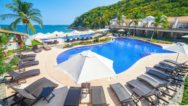A resort pool with lounge chairs and umbrellas, surrounded by palm trees and a view of the ocean.