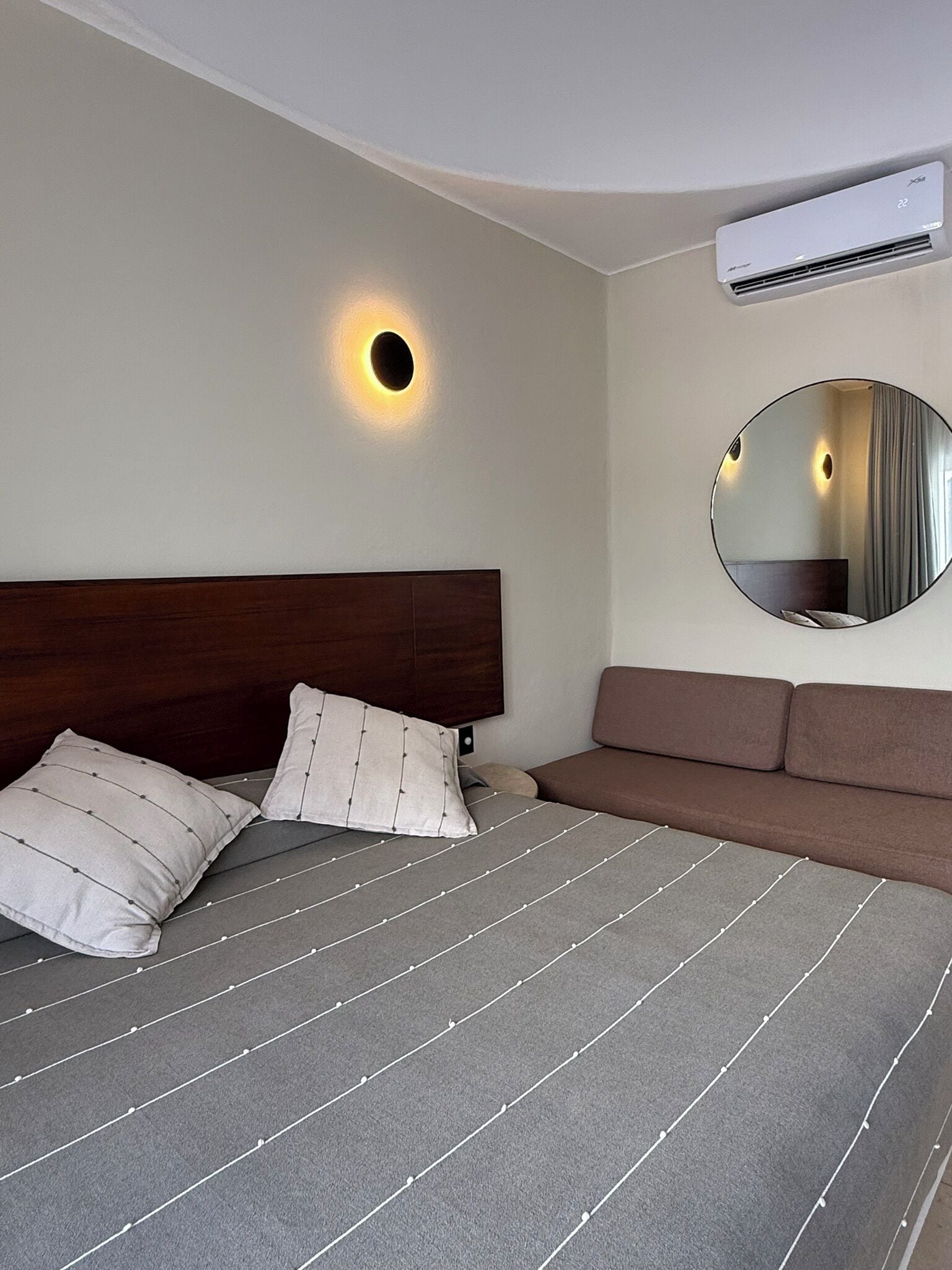 A modern hotel room with a large bed, a headboard, a round mirror, and an air conditioning unit.