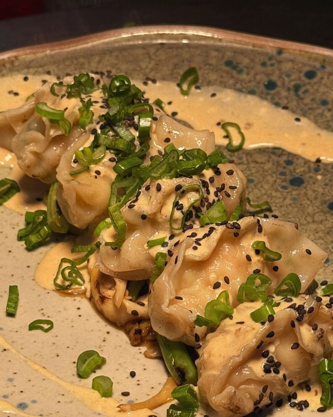 Un plato de dumplings con salsa cremosa, adornado con cebollines y semillas de sésamo negro.