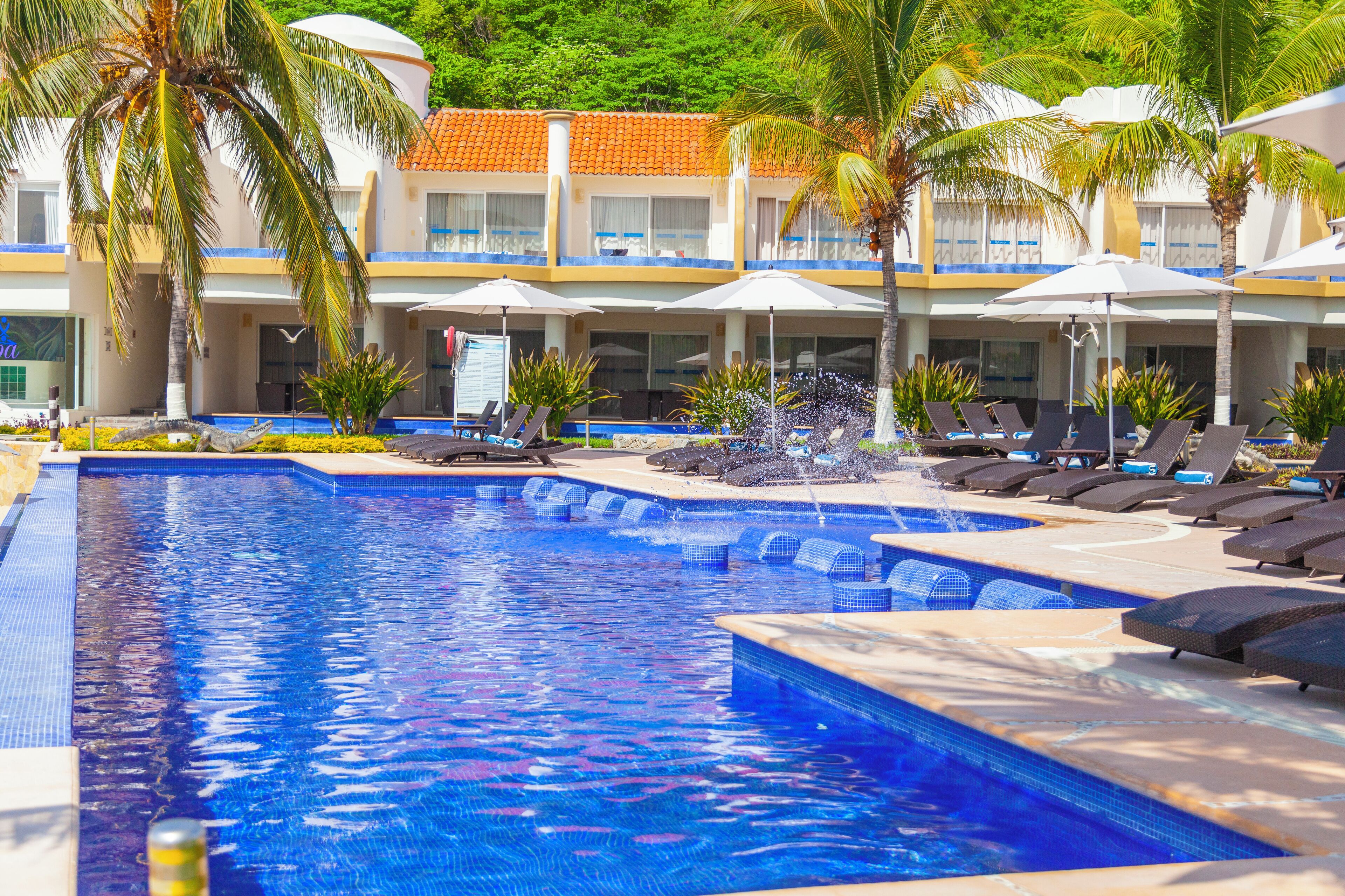A pool area with lounge chairs and umbrellas, surrounded by palm trees and a building with a tiled roof.