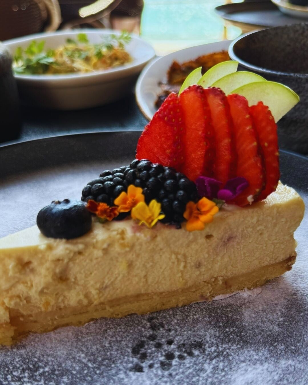 Una porción de pastel de queso cubierta con fresas, zarzamoras, arándanos y flores comestibles.