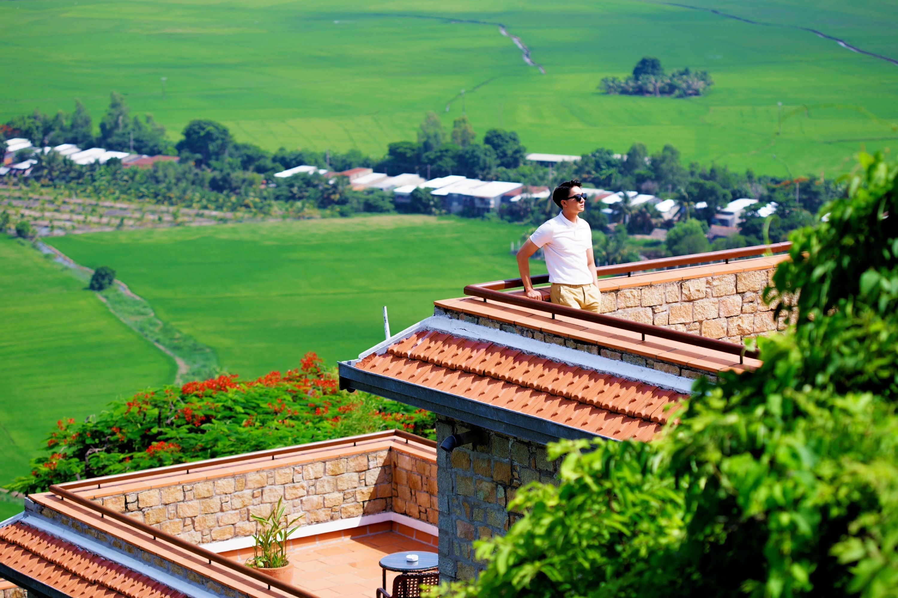 A man standing on a rooftop terrace with a view of green fields and houses.