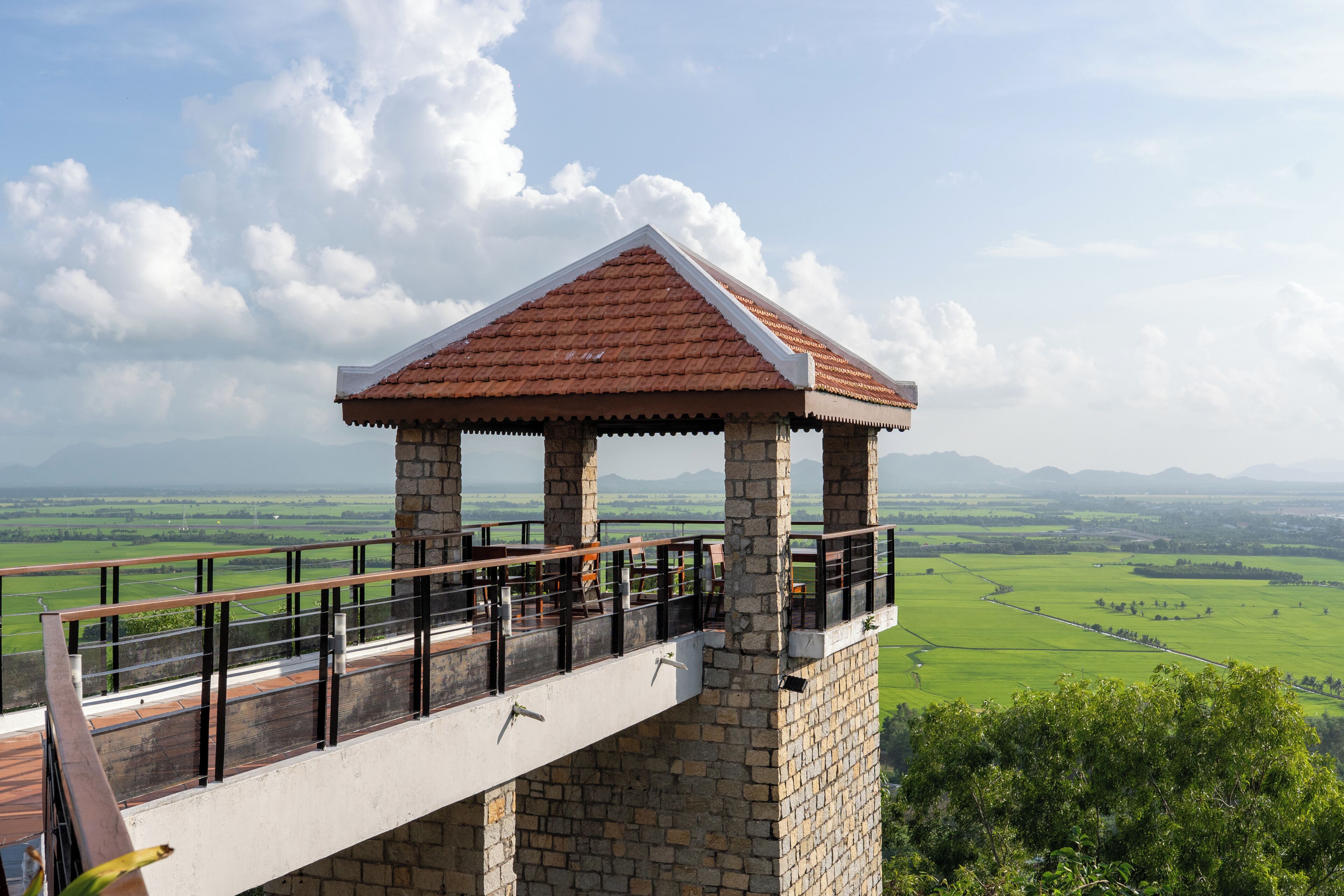 A stone structure with a tiled roof, possibly a lookout or observation point, overlooking a green landscape with fields and distant mountains.