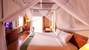 Habitación de hotel con una cama grande, un televisor sobre un escritorio de madera, una silla, una lámpara y vistas al exterior.