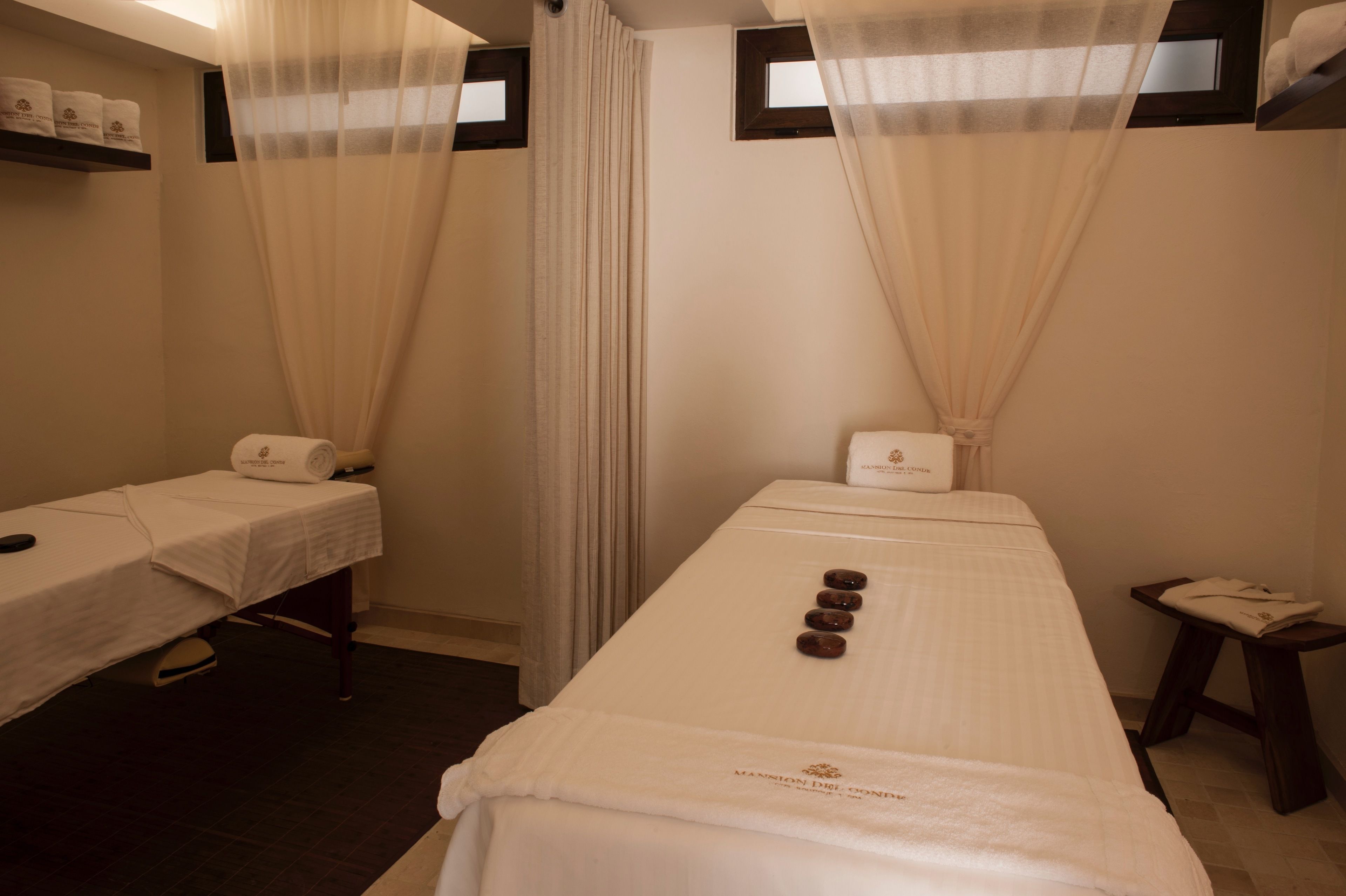 A spa room with two treatment beds, a small table with a towel, and a window with curtains.