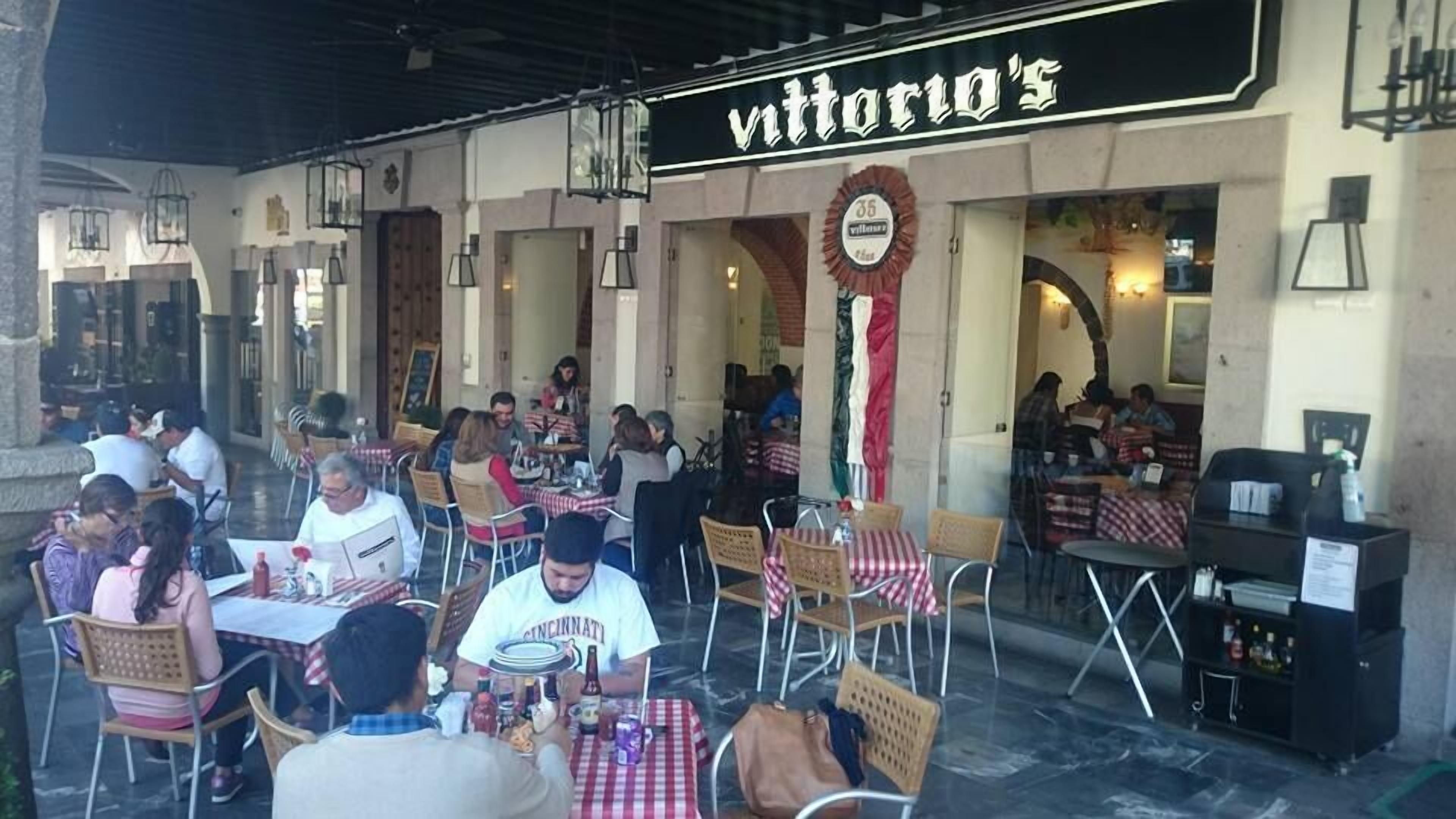 Un espace extérieur aménagé pour les repas, avec des tables et des chaises, une enseigne indiquant « Vittorio's », et une guirlande décorative.