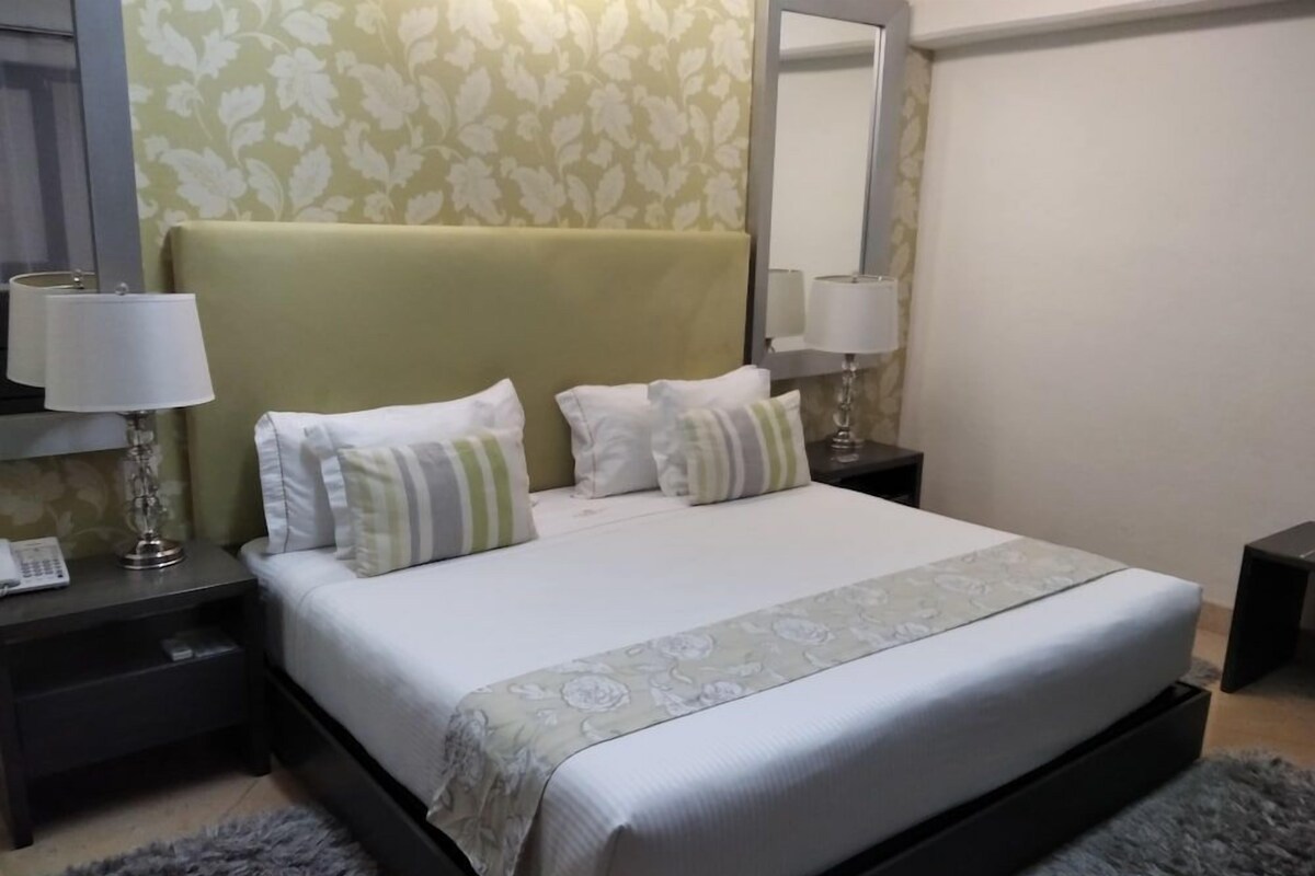 A neatly made bed with a green headboard, white bedding, and striped pillows. There are two bedside tables with lamps and a telephone. The room has a floral wallpaper and a mirror on the wall.