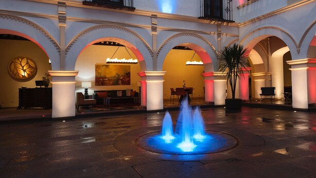 Une cour intérieure dotée d’une fontaine centrale, de colonnes en arc de cercle et d’un espace repas avec des tables et des chaises.