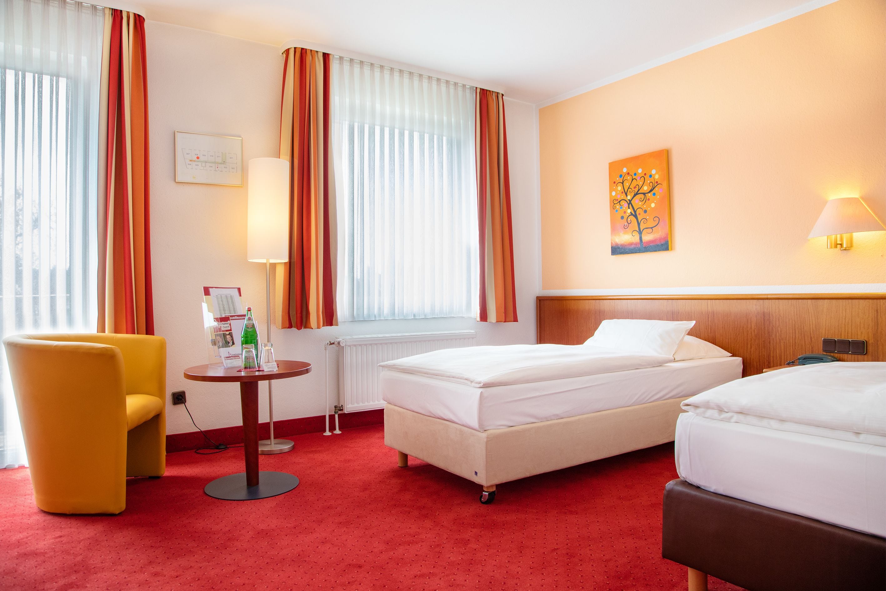 Habitación de hotel con dos camas, una mesita con lámpara, una silla y un cuadro en la pared.