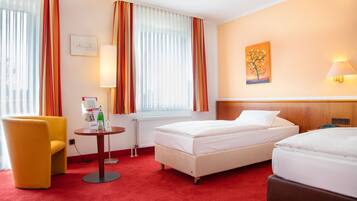 Habitación de hotel con dos camas, una mesita con lámpara, una silla y un cuadro en la pared.
