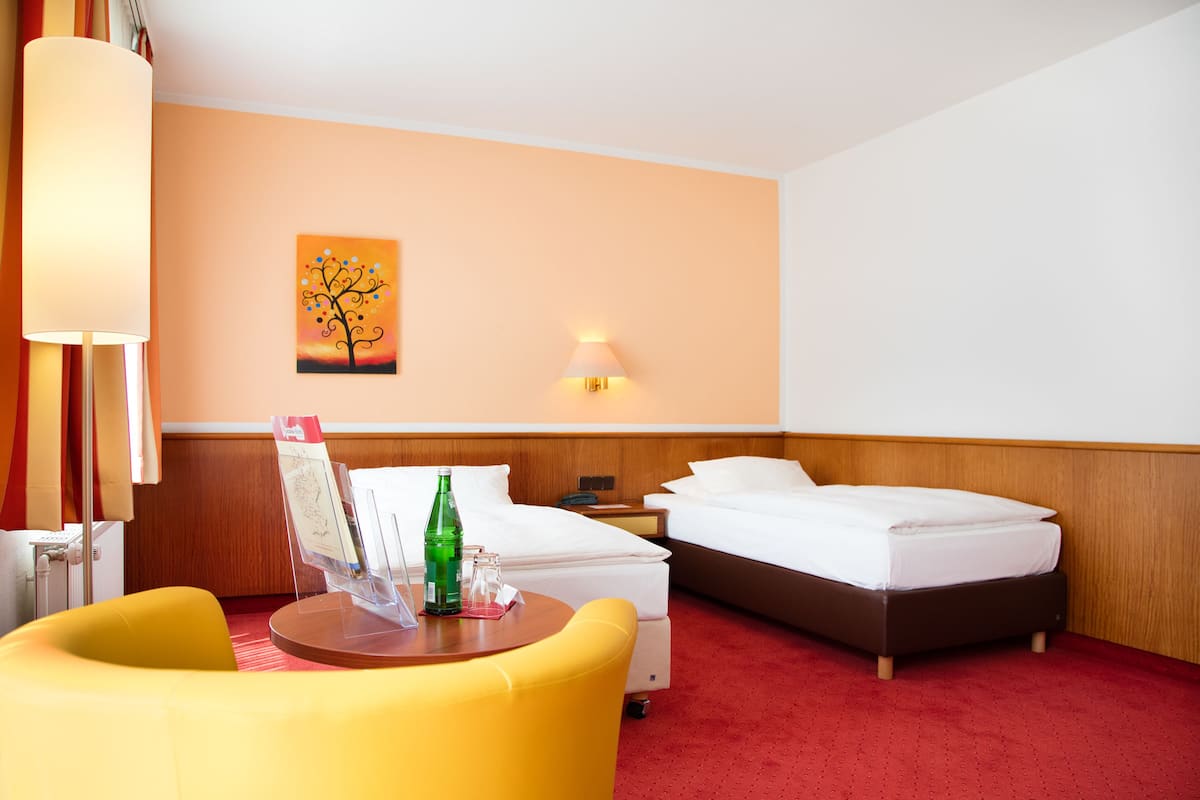A hotel room with two beds, a yellow armchair, a table with a bottle and glasses, a lamp, a wall painting, and a nightstand.