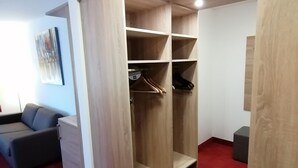 A walk-in closet with wooden shelving and a hanging rod, a gray sofa, and a small gray ottoman. - Montana Hotel Mönchengladbach (Moenchengladbach)