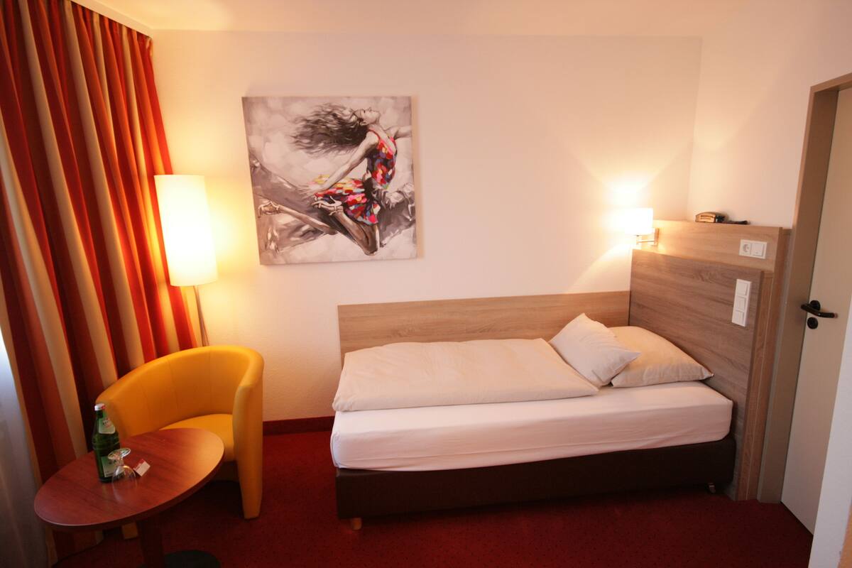 A hotel room with a bed, a chair, a table with a bottle and glass, a painting on the wall, and a lamp.