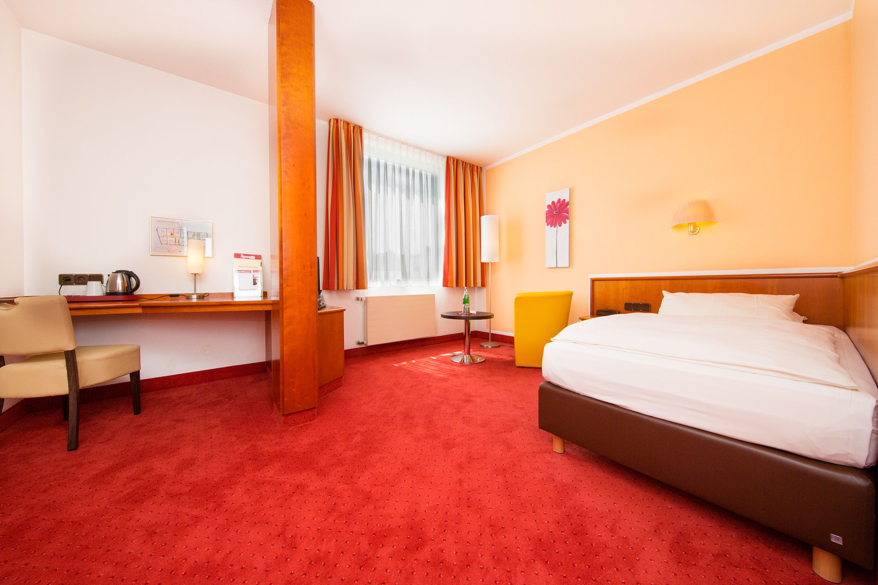 Una camera d'albergo con un letto, una scrivania con una sedia, un tavolino con una lampada e una finestra con tende.