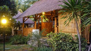 Une construction à toit de chaume, dotée d’une charpente en bois, entourée d’une végétation tropicale et bordée d’un sentier en pierre.