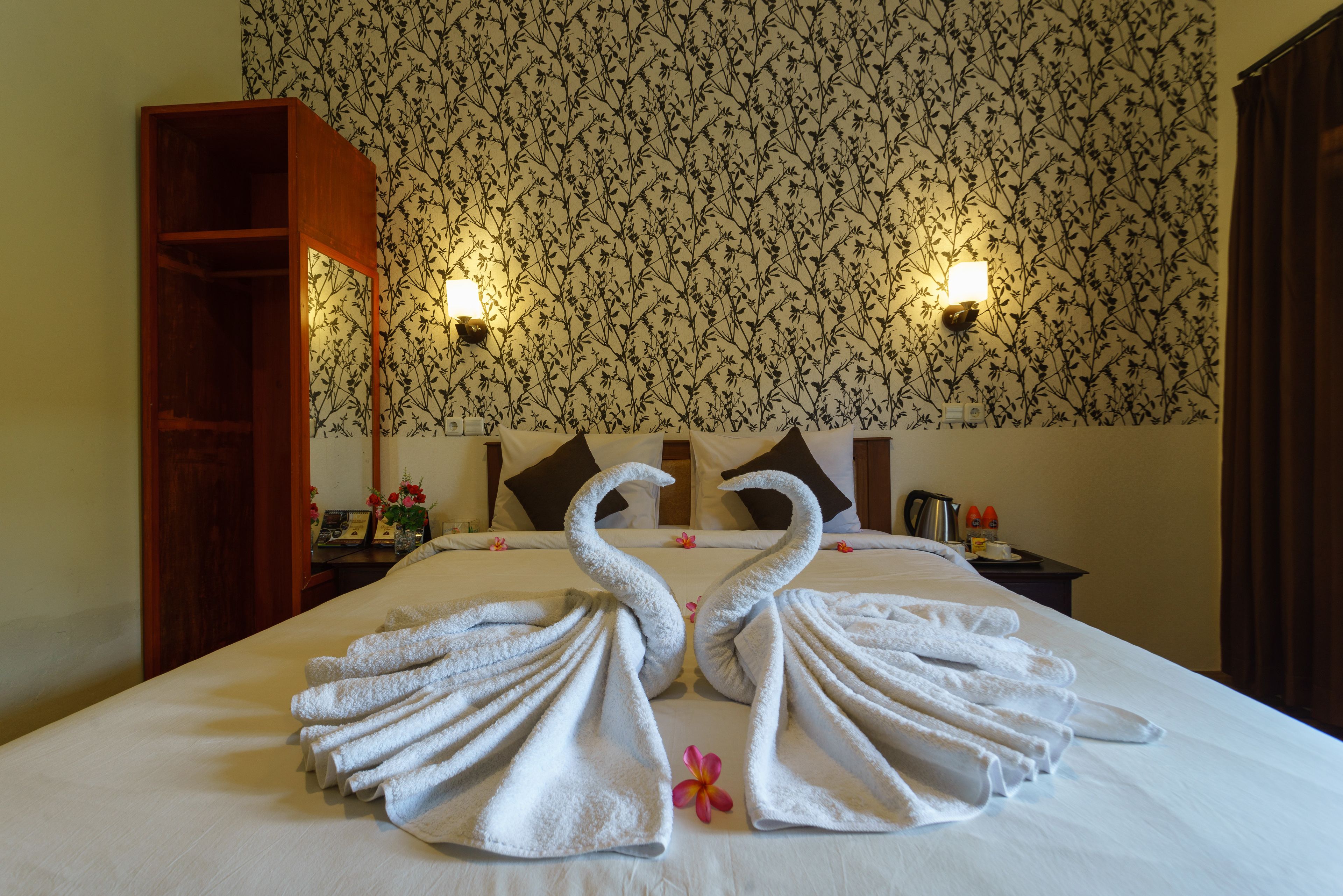 Habitación de hotel con una cama adornada con dos toallas con forma de cisne, papel tapiz floral y dos lámparas de pared.