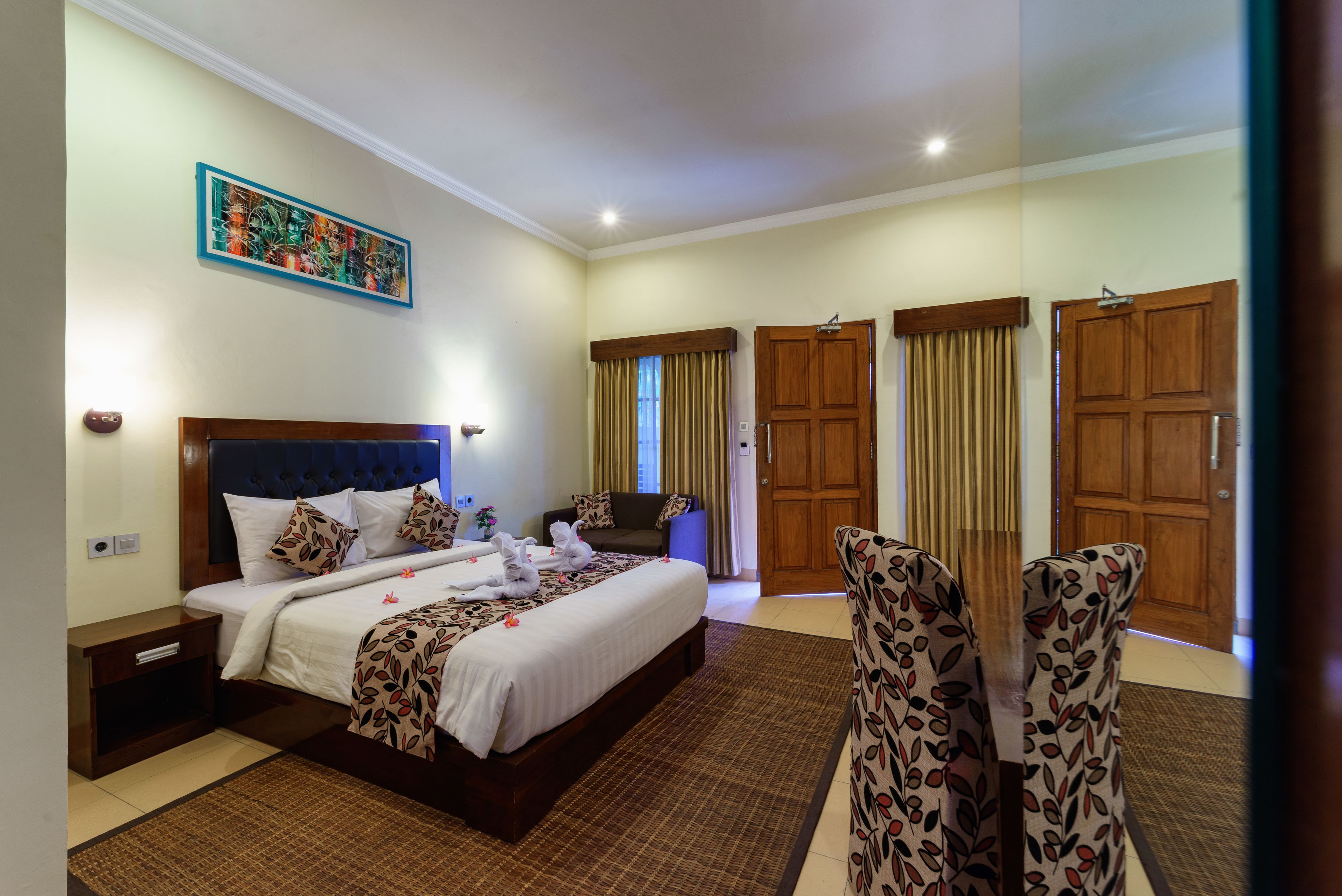 Habitación de hotel con una cama grande, una mesita de noche, un sofá y dos sillas. Hay cortinas en las ventanas y una puerta.