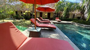 A poolside area with lounge chairs and an umbrella, surrounded by greenery and a building with a tiled roof. - Segara Anak Hotel (Kuta)