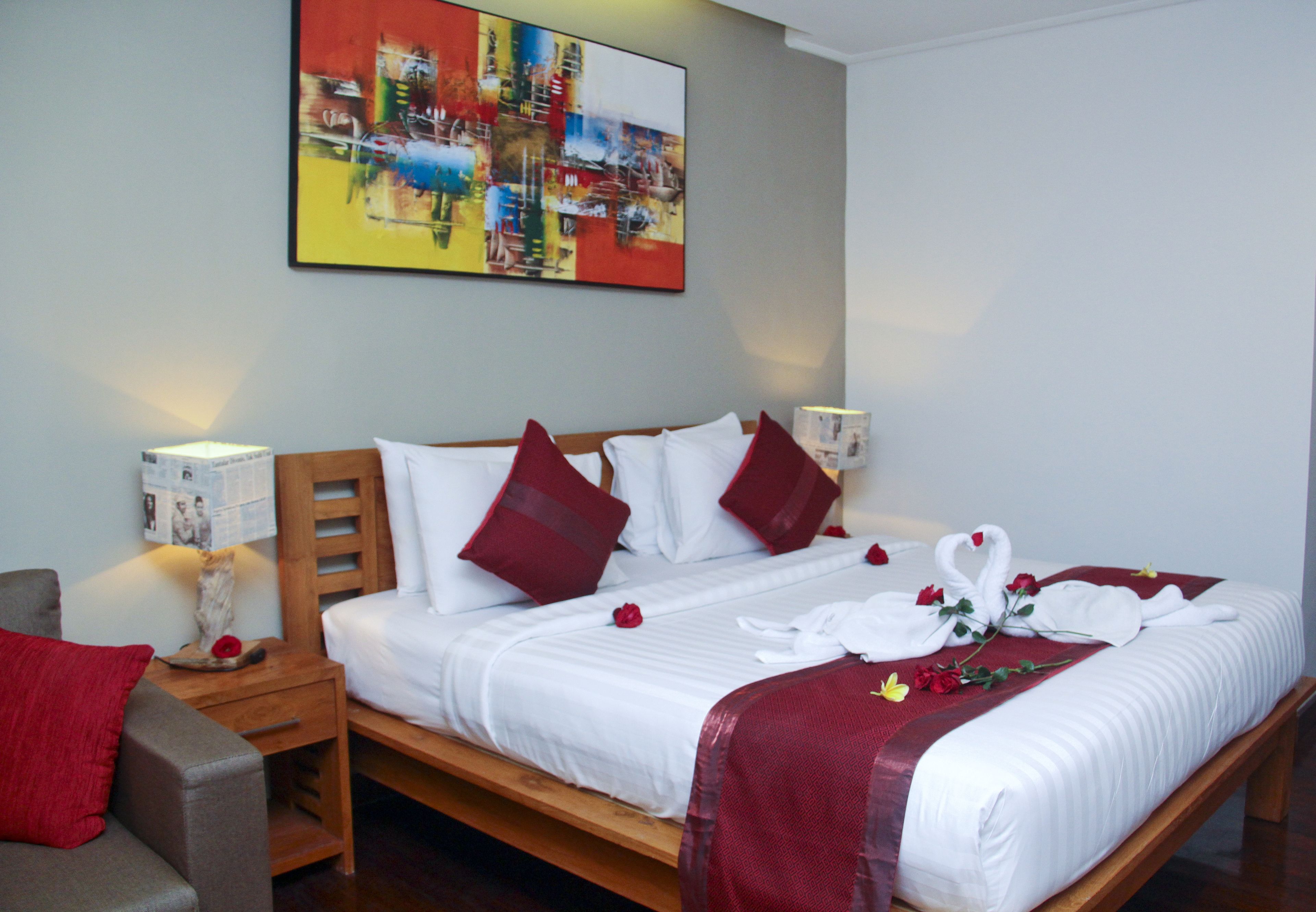 A neatly made bed with white linens and red pillows, a wooden headboard, and a red throw blanket. There is a painting on the wall above the bed and a small table with a lamp beside the bed.
