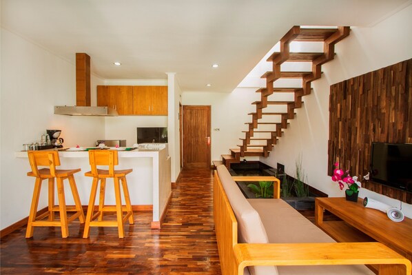 Une cuisine moderne avec des meubles en bois, un îlot central doté de tabourets, un escalier avec des marches en bois et un espace de vie comprenant un canapé et un téléviseur.