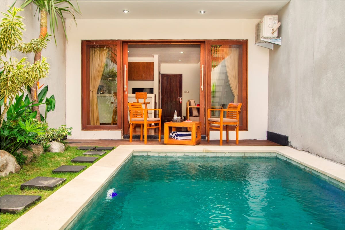 A swimming pool with a wooden deck and chairs, surrounded by a garden with rocks and plants.