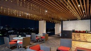 An outdoor seating area with wicker furniture and red cushions, a marble bar, and string lights on the ceiling. - Swiss-Belinn Manyar (Surabaya)
