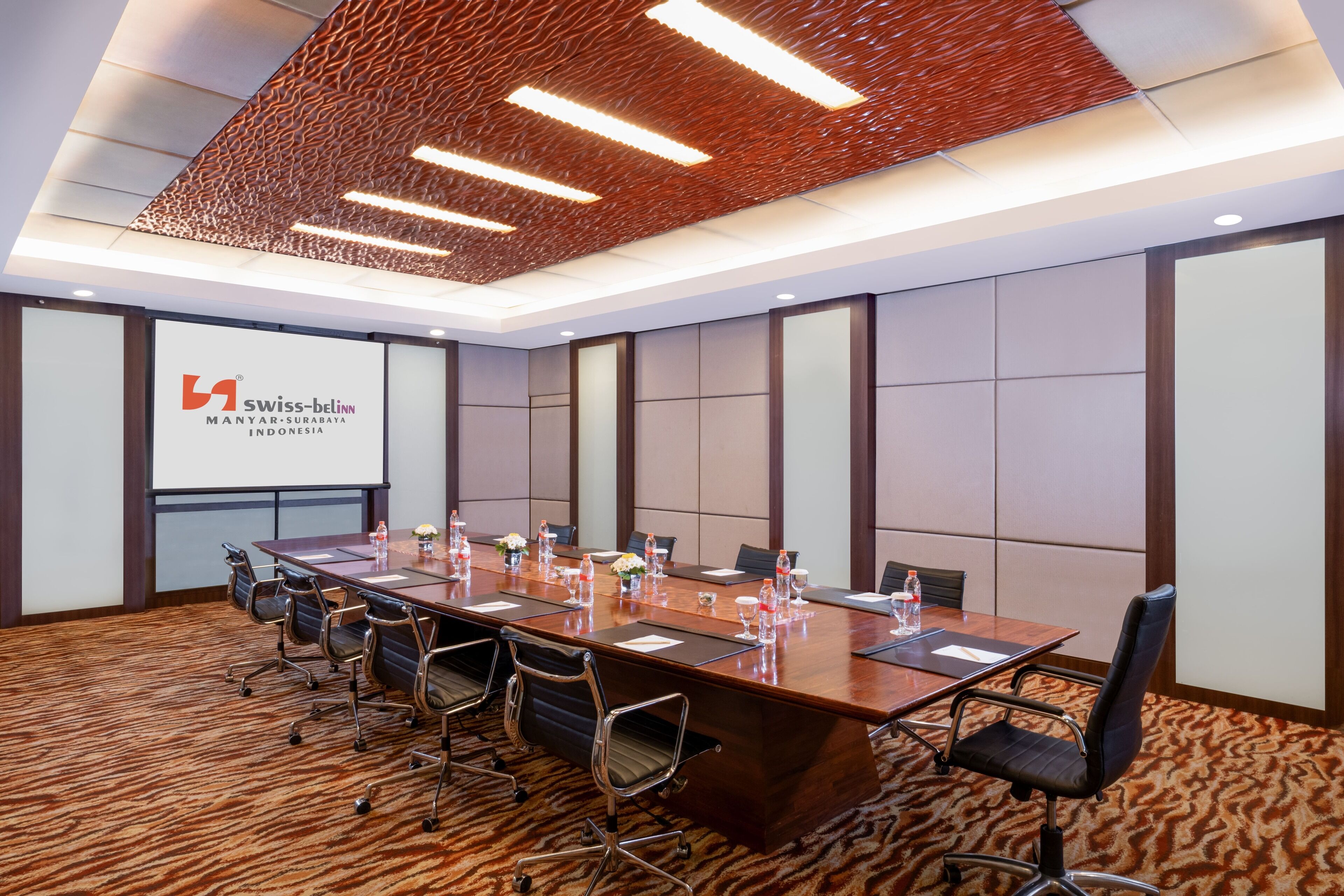 A conference room with a long table, chairs, a projector screen, and a patterned carpet.