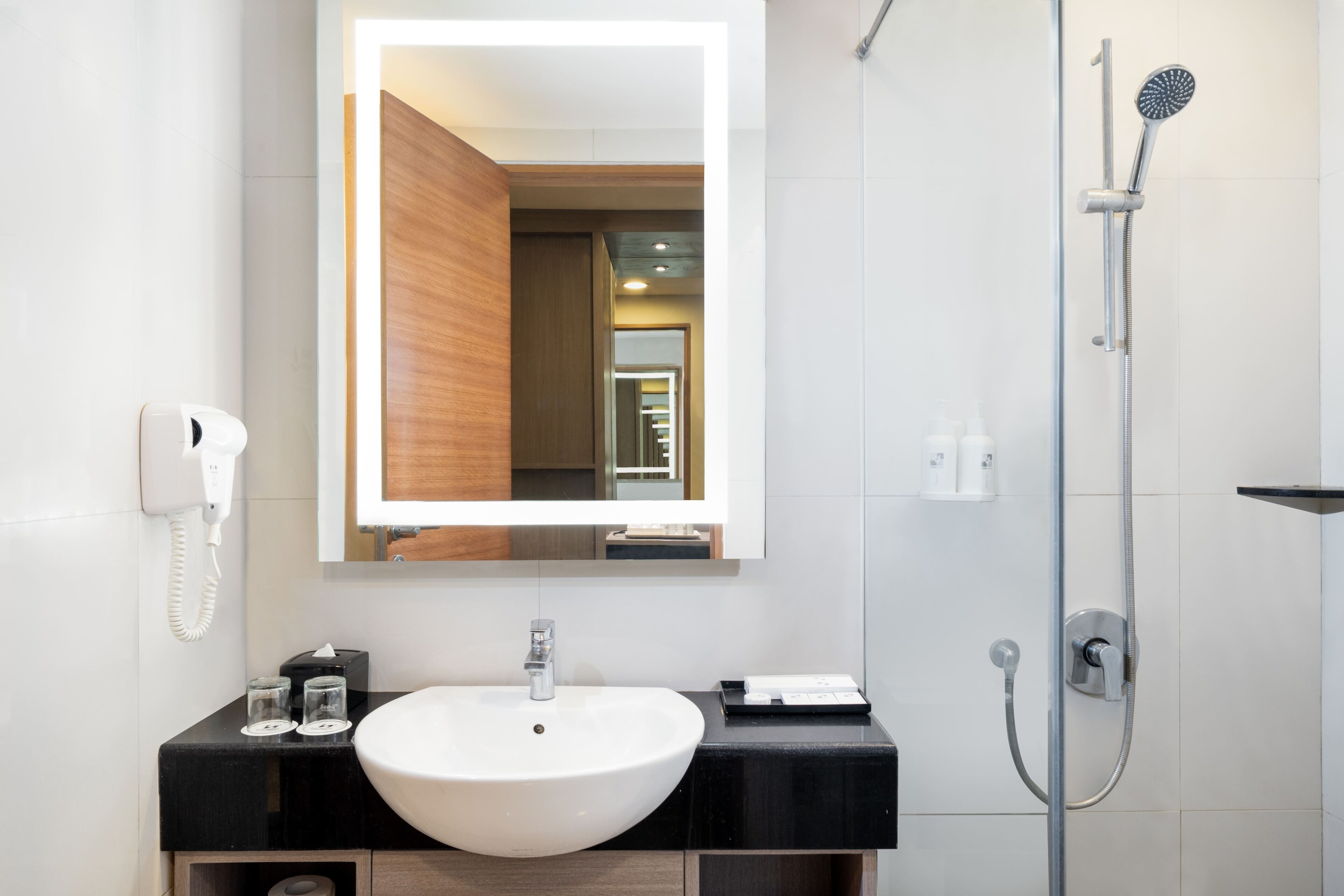 A modern bathroom with a white sink, a mirror with a lighted frame, a handheld showerhead, and a telephone.