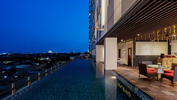 A rooftop infinity pool with a city view, a dining area with chairs and a table, and a bar area with a counter and stools. - Swiss-Belinn Manyar (Surabaya)
