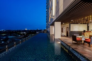 Una piscina infinita en la azotea con vistas a la ciudad, una zona de comedor con sillas y una mesa, y un bar con mostrador y taburetes.