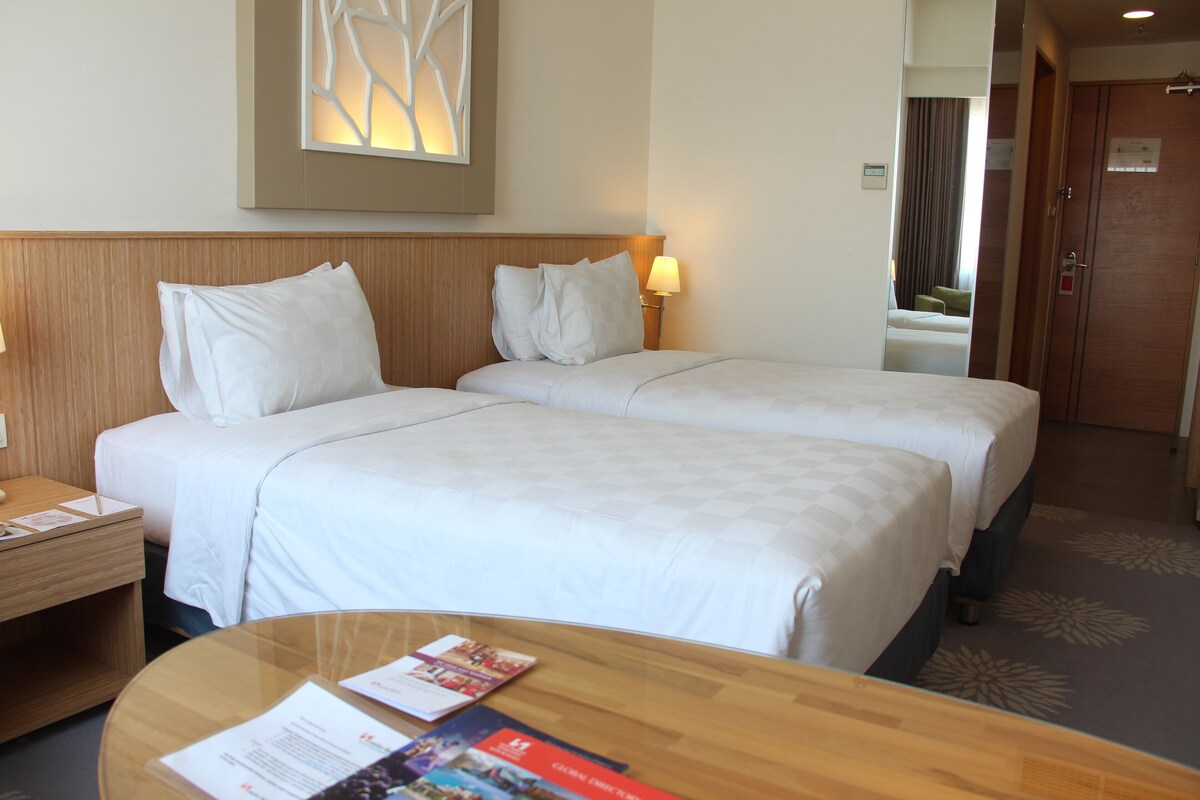A hotel room with two beds, a nightstand, a lamp, a glass table with brochures, and a wall-mounted artwork.