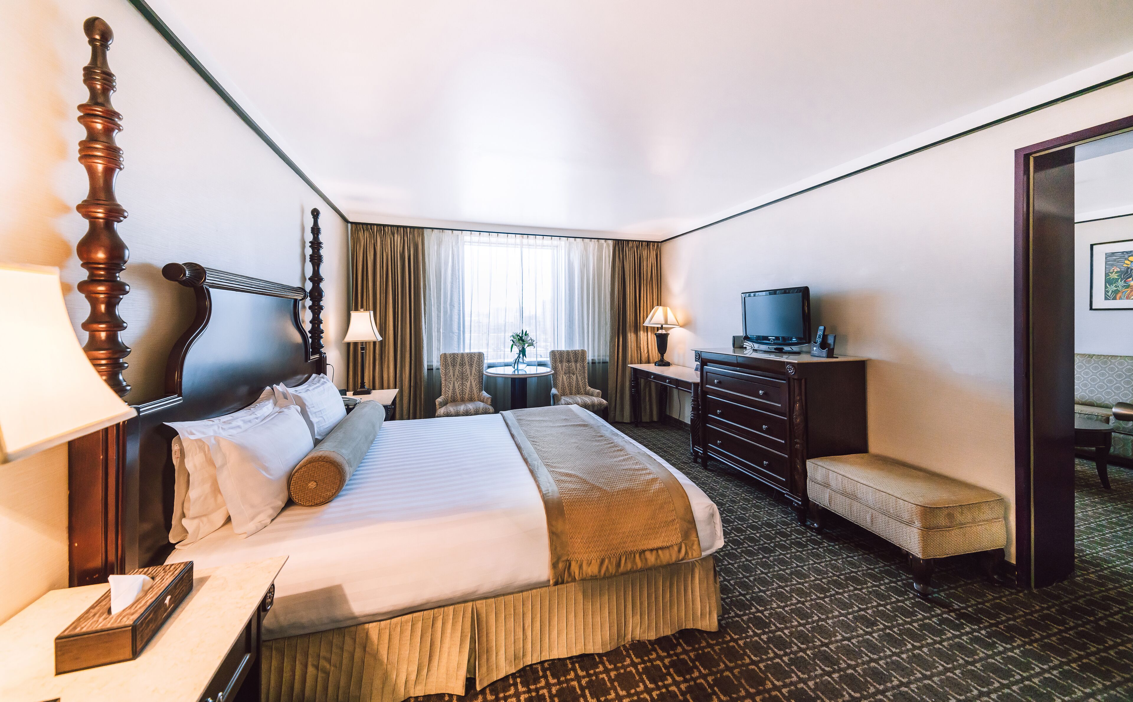 A hotel room with a large bed, a desk with a television, a chair, a lamp, and a small table with a plant.