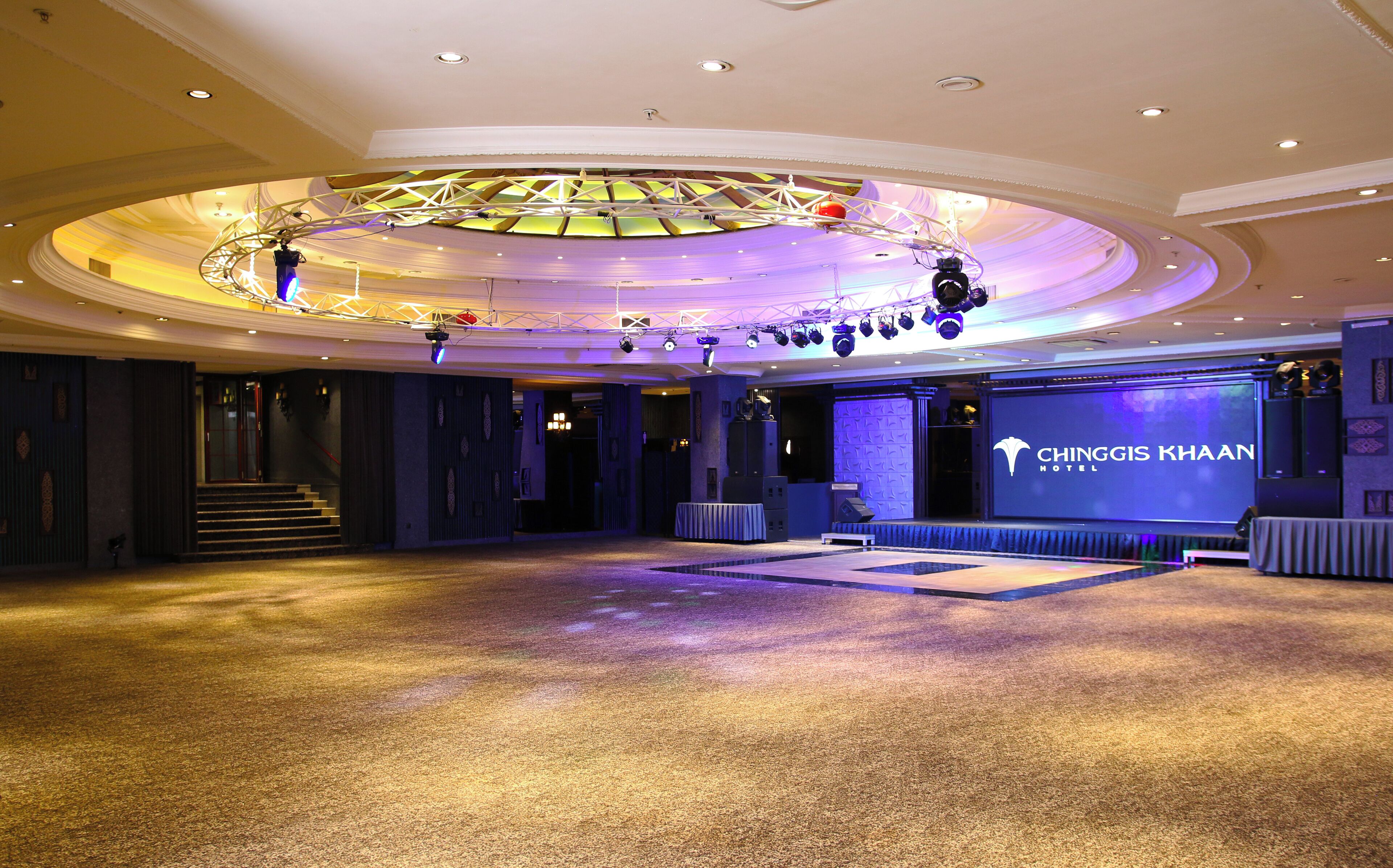 A spacious hotel lobby with a stage, a large screen displaying the hotel name, and a grand chandelier.