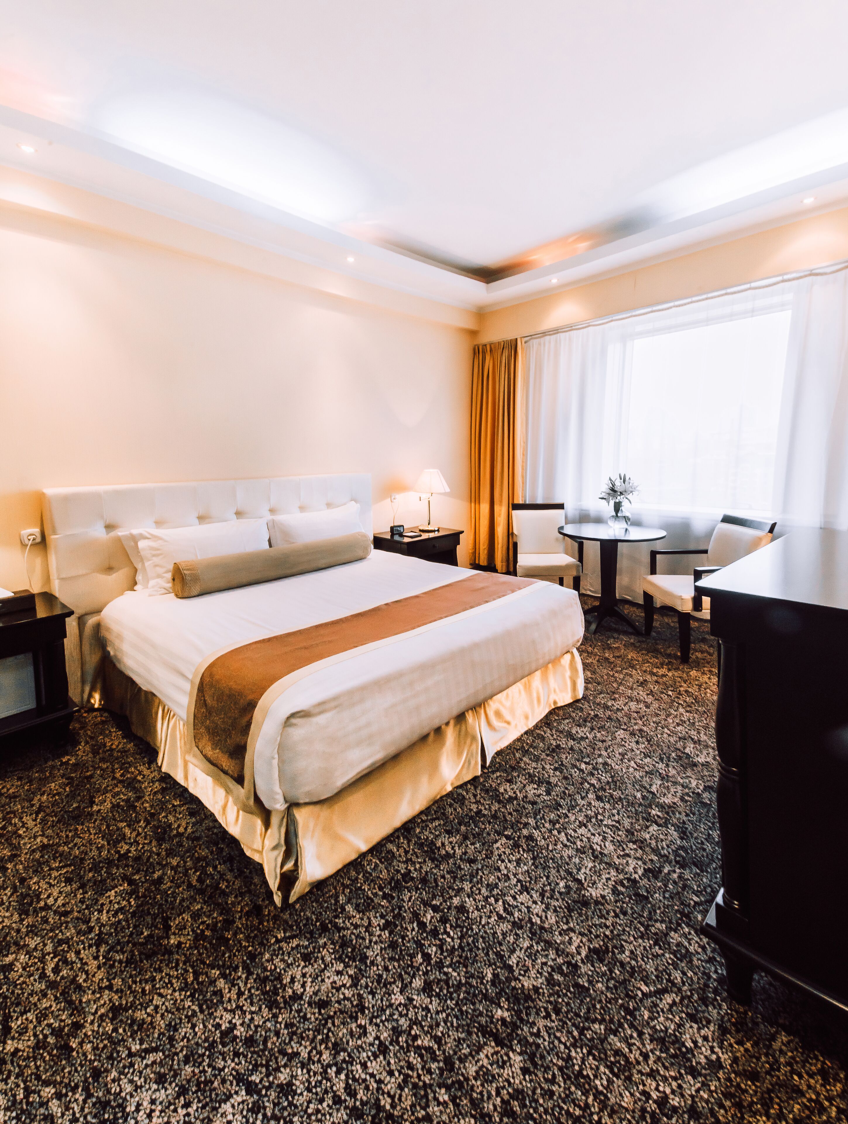 A hotel room with a large bed, a desk, a chair, a small table, and a window with curtains.