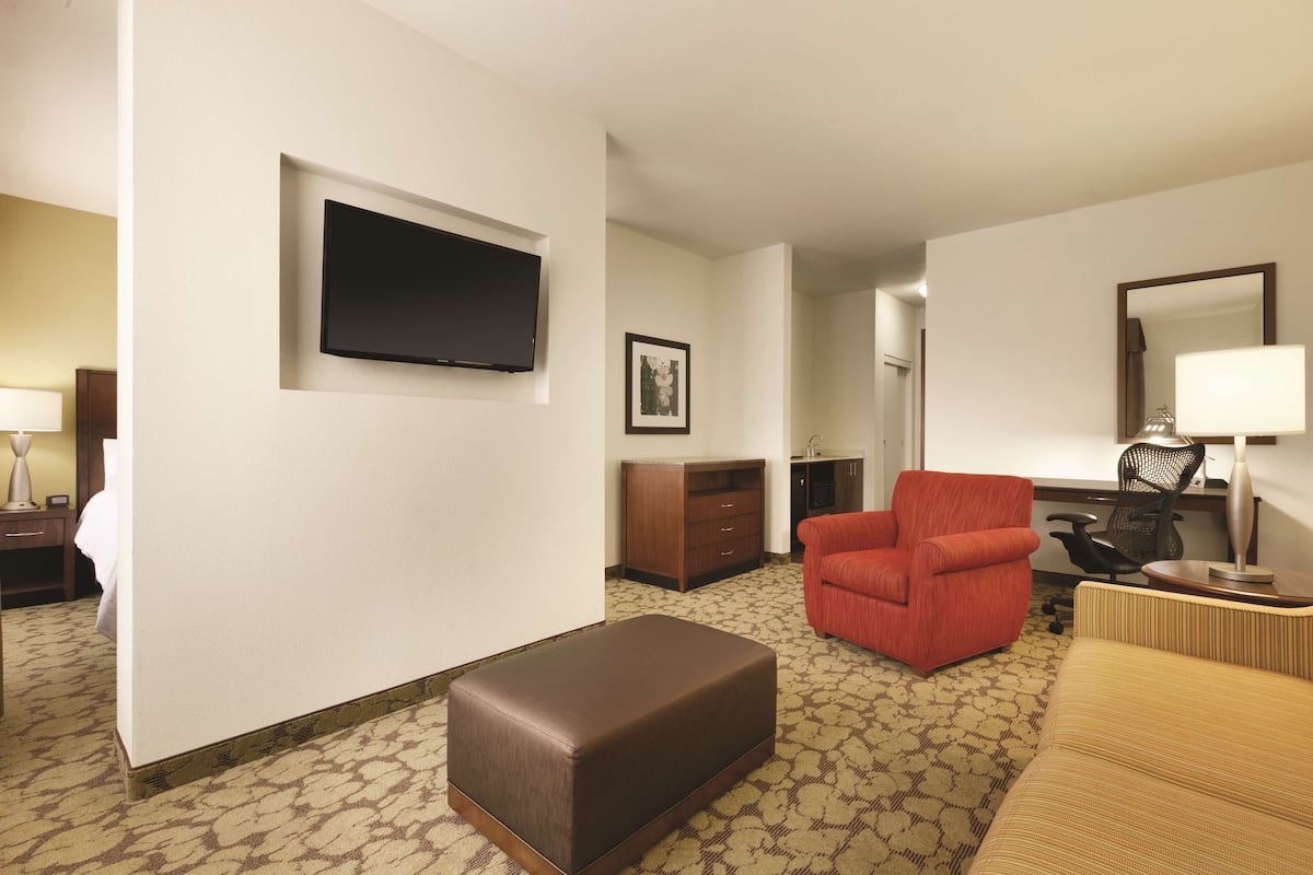 A hotel room with a flat-screen TV mounted on the wall, a brown ottoman, a red armchair, a sofa, a desk with a chair, a lamp, and a framed picture on the wall.