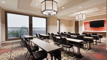 A conference room with tables and chairs, large windows with a view of the water, and a flat-screen TV mounted on the wall.