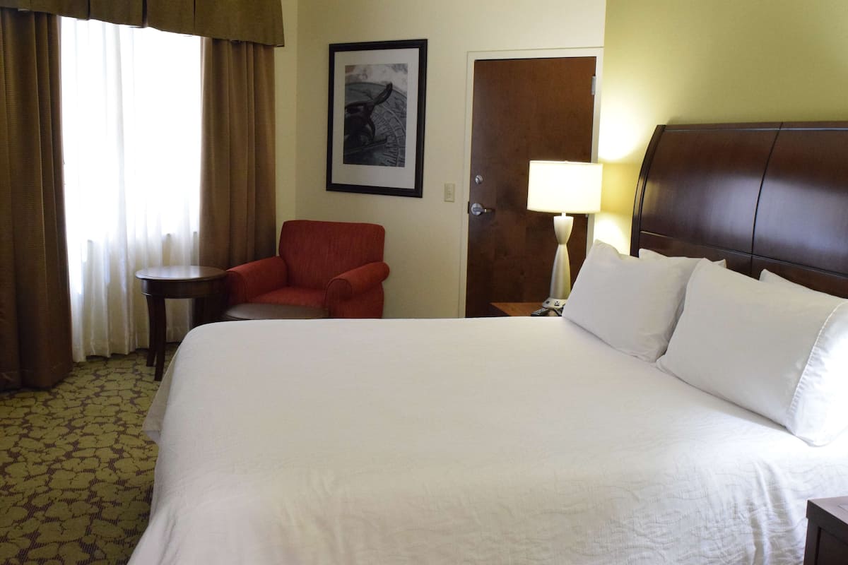 A hotel room with a large bed, a red armchair, a small table, a framed picture on the wall, and a door with a nightstand and lamp.