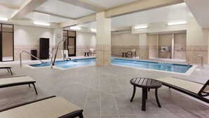 An indoor swimming pool area with multiple lounge chairs and a small table, surrounded by tiled flooring and walls.