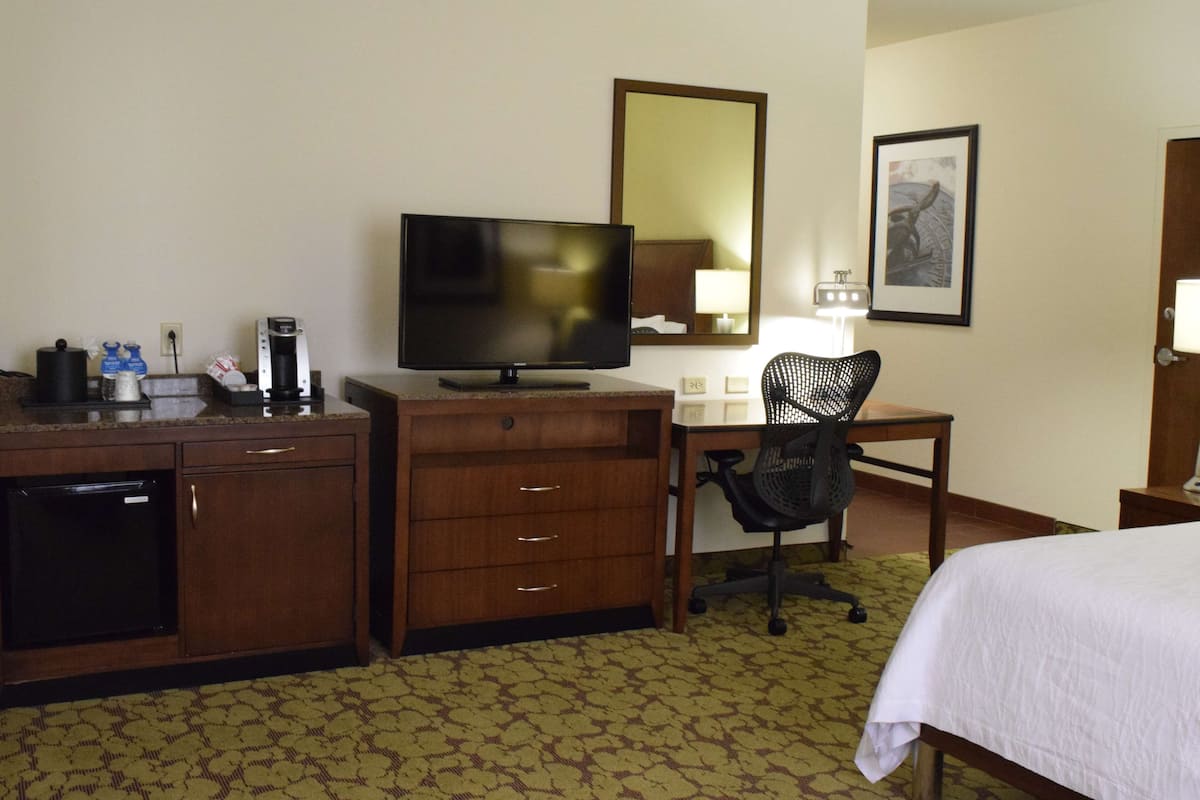 A hotel room with a bed, a desk with a computer, a chair, a television, a coffee maker, and a minibar.