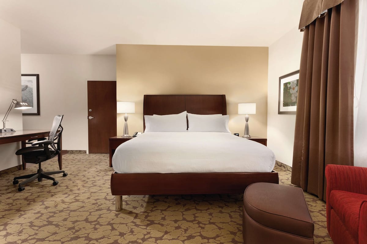 A hotel room with a large bed, a desk with a chair, a lamp, and a red armchair.