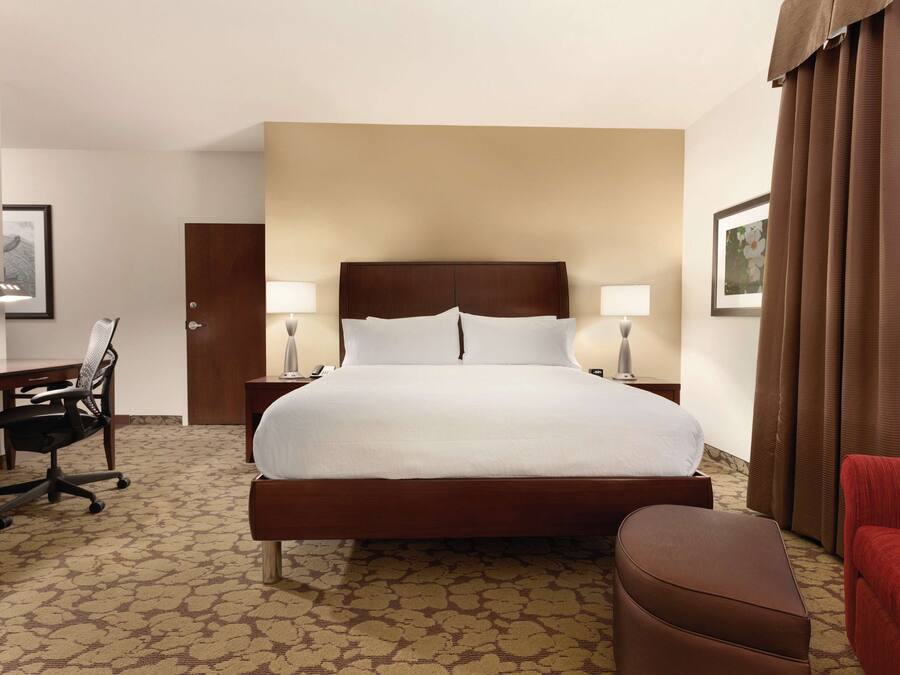 A hotel room with a large bed, a desk with a chair, a lamp, and a red armchair.