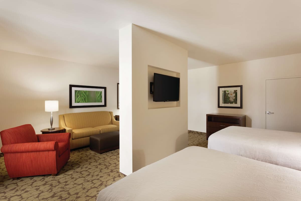 A hotel room with two beds, a red armchair, a yellow sofa, a flat-screen TV mounted on the wall, and a framed picture above the sofa.